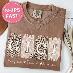 Pode incluir: Camiseta cor espresso com a palavra "GIGI" em um design &uacute;nico. As letras est&atilde;o em um fundo de estampa de leopardo e xadrez. Os nomes Megan, Sarah e Lauren s&atilde;o impressos abaixo. Um c&iacute;rculo rosa com "SHIPS FAST!" &eacute; vis&iacute;vel.