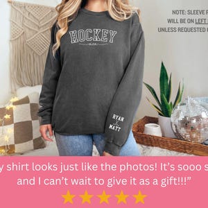 K&ouml;nnte beinhalten: Ein dunkelgrauer Pullover mit dem Wort "HOCKEY" auf der Vorderseite und den Namen "RYAN & MATT" auf dem linken &Auml;rmel. Der Pullover wird von einer Person getragen.