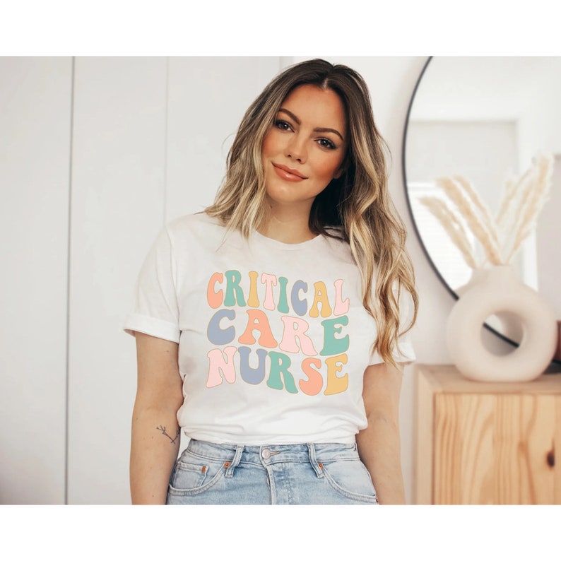 Peut inclure: Un t-shirt blanc avec le texte "CRITICAL CARE NURSE" en lettres r&eacute;tro color&eacute;es.