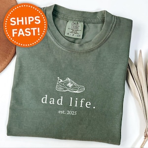 May include: A green t-shirt with a white graphic of a sneaker and the text "dad life. est. 2025".