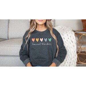 May include: A dark gray sweatshirt with a row of six colorful hearts and the text "Social Worker" in white cursive writing.