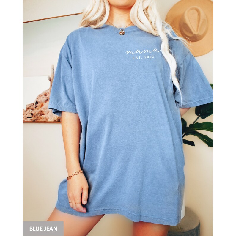 Pu&ograve; includere: Una t-shirt oversize in jeans blu con il testo "mama est. 2023" in bianco.