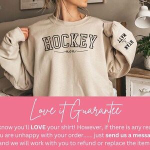 K&ouml;nnte beinhalten: Ein beiger Pullover mit dem Aufdruck "HOCKEY MOM" auf der Vorderseite. Die &Auml;rmel haben die Namen "MATT" und "RYAN" aufgedruckt.