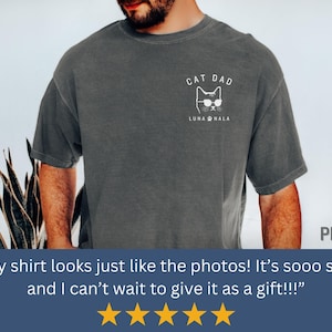 May include: Dark gray t-shirt with a white graphic of a cat wearing sunglasses and the text "CAT DAD" and "LUNA NALA". The shirt is a comfortable, casual style.