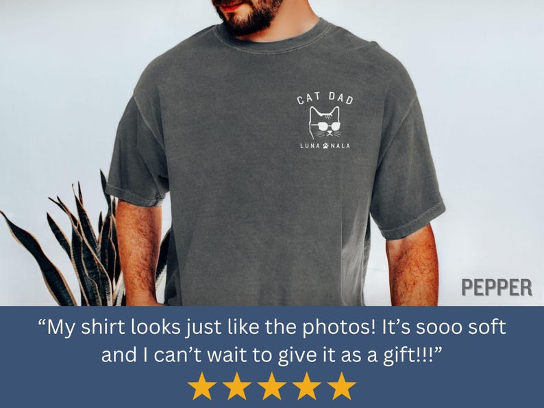 May include: Dark gray t-shirt with a white graphic of a cat wearing sunglasses and the text "CAT DAD" and "LUNA NALA". The shirt is a comfortable, casual style.