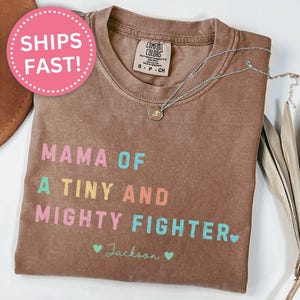 May include: Espresso-colored t-shirt with the text "MAMA OF A TINY AND MIGHTY FIGHTER" in colorful letters. A silver necklace with a pendant rests on the shirt. The name "Jackson" is below the text, with two small hearts.