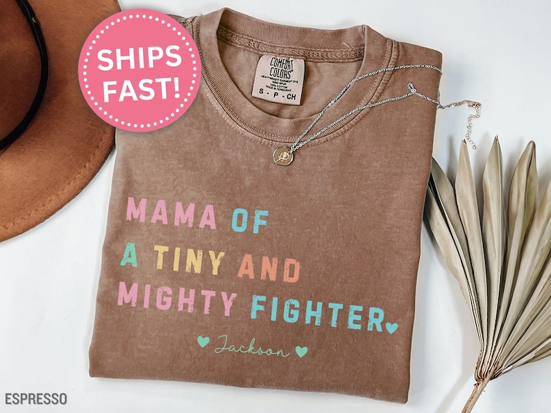 May include: Espresso-colored t-shirt with the text "MAMA OF A TINY AND MIGHTY FIGHTER" in colorful letters. A silver necklace with a pendant rests on the shirt. The name "Jackson" is below the text, with two small hearts.