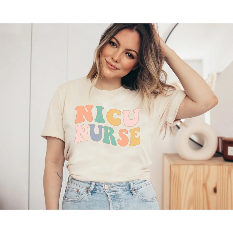 Pode incluir: Uma mulher vestindo uma camiseta bege claro com o texto "NICU NURSE" em letras retr&ocirc; coloridas. A camiseta tem mangas curtas e gola redonda.