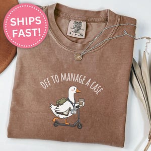 May include: Espresso-colored t-shirt with the phrase "OFF TO MANAGE A CASE" and a cartoon goose riding a scooter, holding a coffee cup. The shirt has a Comfort Colors label and a small silver necklace.
