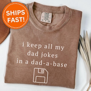 May include: Espresso-colored t-shirt with the phrase "i keep all my dad jokes in a dad-a-base" in white text. The design includes a floppy disk graphic. The shirt has a Comfort Colors label.