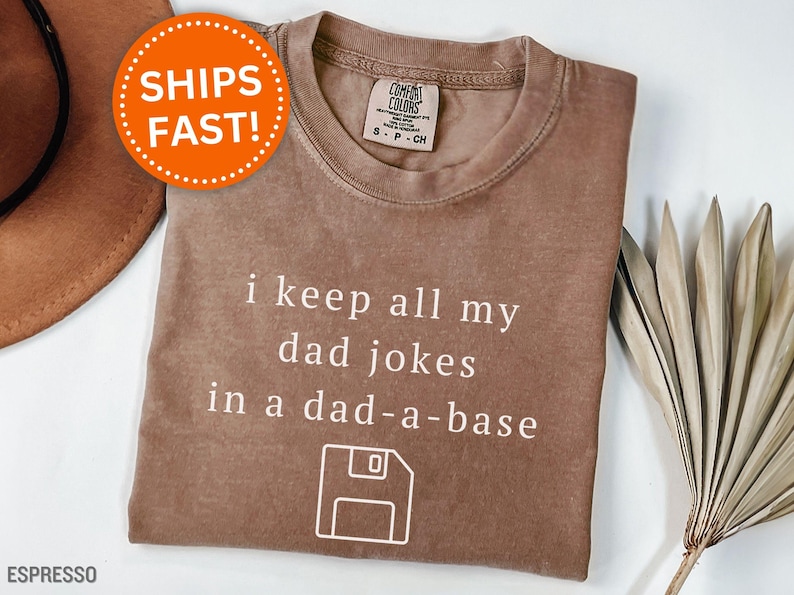 May include: Espresso-colored t-shirt with the phrase "i keep all my dad jokes in a dad-a-base" in white text. The design includes a floppy disk graphic. The shirt has a Comfort Colors label.