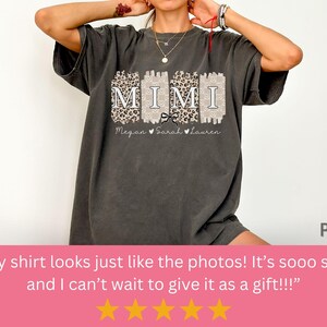Może przedstawiać: Szary t-shirt z nadrukiem graficznym słowa "MIMI" białymi literami z czarną kokardką. Grafika jest otoczona wzorem w panterkę. Tekst "Megan Sarah Lauren" jest nadrukowany pod grafiką.