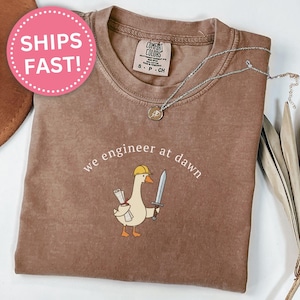 May include: An espresso-colored t-shirt featuring a graphic of a goose wearing a yellow hard hat, holding a rolled-up blueprint in one wing and a sword in the other, with the text "we engineer at dawn" printed in white above the goose illustration.