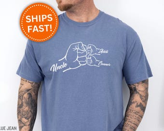 Puede incluir: Camiseta azul vaquero con un gráfico blanco de tres puños chocando. La palabra "Uncle" está encima del puño más grande, y los nombres "Addi" y "Connor" están encima de los puños más pequeños. Un círculo naranja con las palabras "SHIPS FAST!" está en la parte superior izquierda.