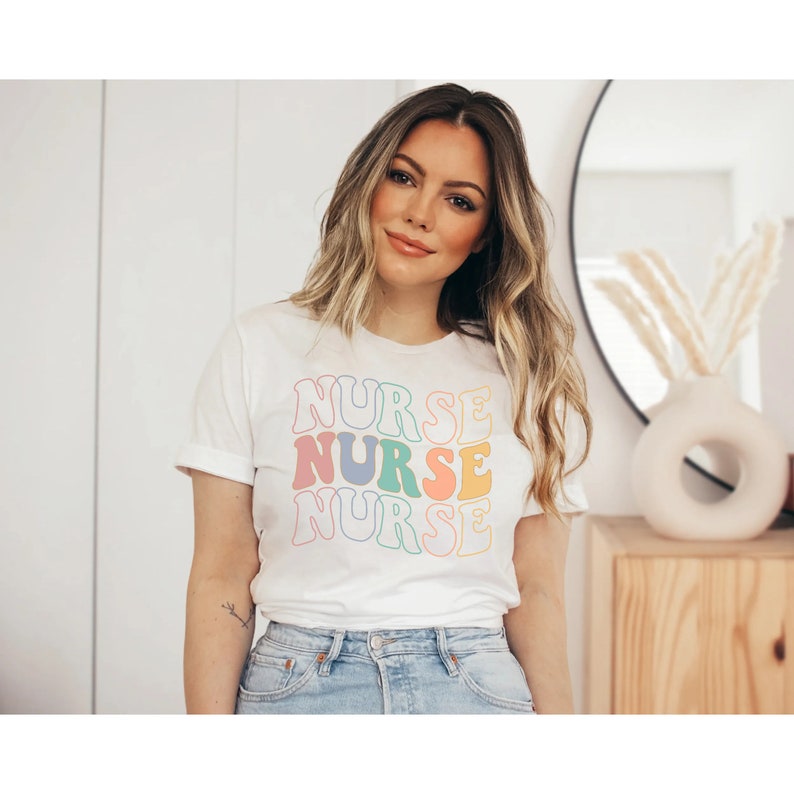 Pu&ograve; includere: Maglietta bianca con la parola "NURSE" ripetuta tre volte in uno schema di colori arcobaleno. La parola &egrave; in uno stile di font retr&ograve;.