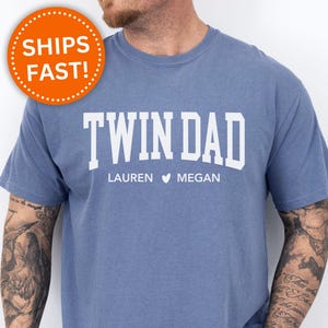 May include: A blue jean t-shirt with the text "Twin Dad" in white letters. Below the text, it says "Lauren Megan" with a heart symbol between the names.