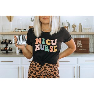 Pode incluir: Uma camiseta preta com o texto "NICU NURSE" em letras retr&ocirc; coloridas. As letras s&atilde;o rosa, amarelo, azul e verde. A pessoa que est&aacute; usando a camiseta est&aacute; segurando uma caneca de prata.