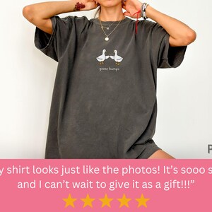 May include: Dark gray t-shirt featuring a graphic of two white geese and the text "goose bumps." The shirt has a relaxed fit and is made of a soft material. The shirt is part of a clothing collection.
