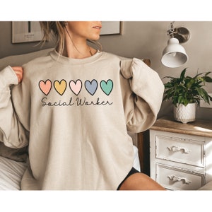 May include: A beige sweatshirt with a row of five colorful hearts and the text "Social Worker" in cursive.