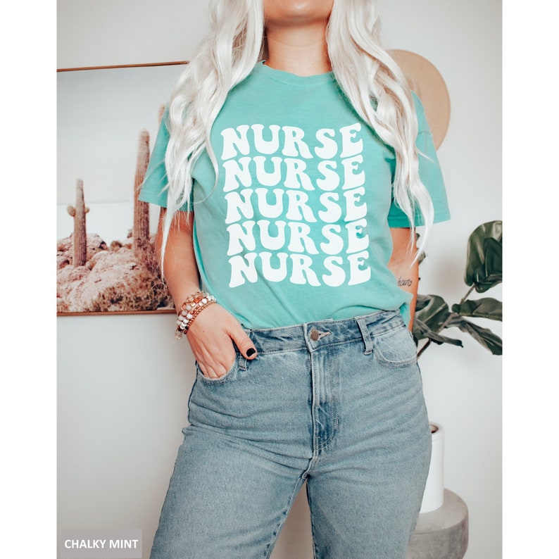 May include: A turquoise blue t-shirt with the word "NURSE" repeated five times in a retro font. The shirt is being worn by a person with blonde hair.