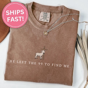 May include: Espresso-colored t-shirt with the text "HE LEFT THE 99 TO FIND ME" and a graphic of a lamb and cross. A silver necklace with a pendant rests on the shirt. A pink sticker with the words "SHIPS FAST!" is visible.