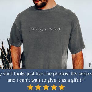 May include: A dark gray t-shirt with the text "hi hungry, i'm dad." printed in white on the front.