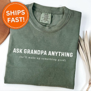 May include: Moss green t-shirt with the text "ASK GRANDPA ANYTHING (he'll make up something good)". The shirt has a crew neck and short sleeves. A brown hat and dried plant are in the background.