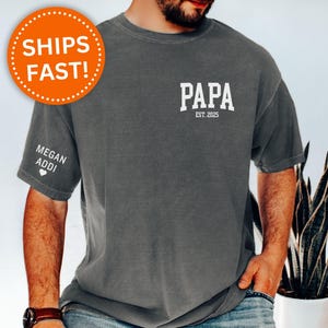 Op de afbeelding: Donkergrijs t-shirt met het woord "PAPA" en "EST. 2025" in het wit op de voorkant. De linkermouw heeft de tekst "MEGAN ADDI" en een hart. Het shirt heeft een relaxte pasvorm.