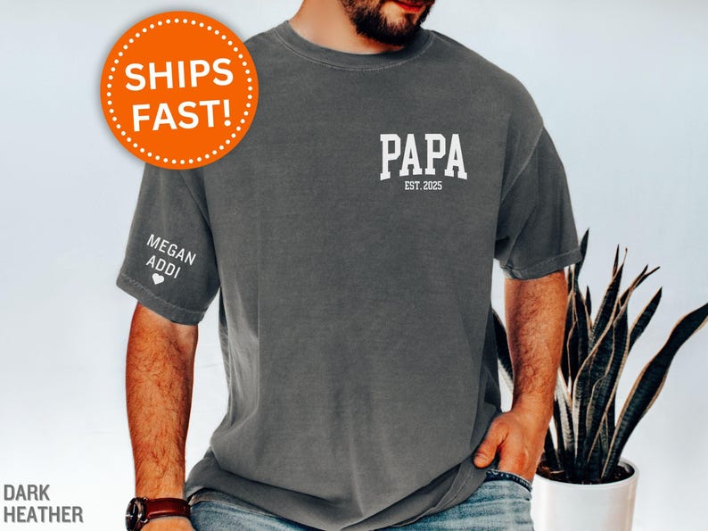 Op de afbeelding: Donkergrijs t-shirt met het woord "PAPA" en "EST. 2025" in het wit op de voorkant. De linkermouw heeft de tekst "MEGAN ADDI" en een hart. Het shirt heeft een relaxte pasvorm.