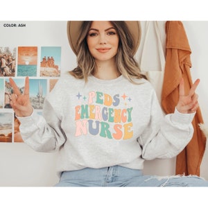 May include: A gray sweatshirt with the text "PEDS EMERGENCY NURSE" in colorful retro-style lettering. The text is surrounded by small white stars.