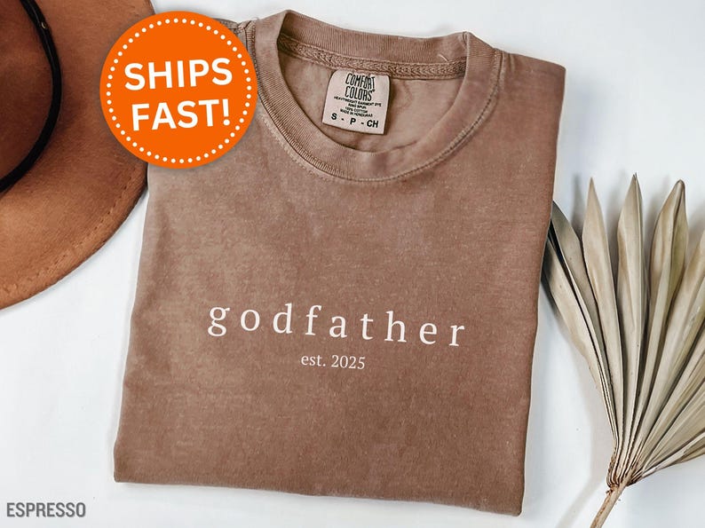 May include: Espresso-colored t-shirt with the word "godfather" and "est. 2025" printed in white. The shirt has a crew neck and a Comfort Colors tag. A round orange sticker with the words "SHIPS FAST!" is visible.