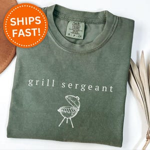 Op de afbeelding: Mosgroen t-shirt met de woorden "grill sergeant" en een grillafbeelding. Het shirt heeft een ronde hals en een klein label op de rug. Een bruine hoed en gedroogde bladeren zijn op de achtergrond.