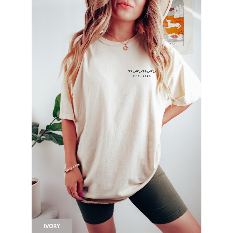 Pu&ograve; includere: Una donna indossa una maglietta color crema con la scritta "mama est. 2023" sul davanti. Indossa anche pantaloncini da ciclista verde scuro.