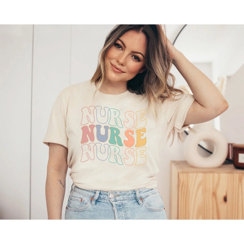 Pu&ograve; includere: Una donna indossa una maglietta beige chiaro con la parola "NURSE" ripetuta tre volte in uno schema di colori arcobaleno. Il testo &egrave; in un font retr&ograve; e ondulato.