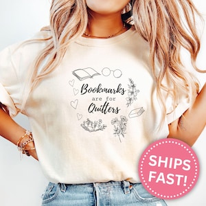 Può includere: T-shirt color avorio con un design circolare che include un libro aperto, occhiali, cuori e fiori. Il testo "Bookmarks are for Quitters" è al centro. Un cerchio rosa con "SHIPS FAST!" è in basso a destra.