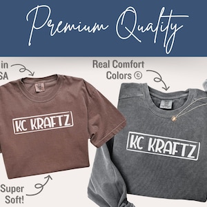 May include: Two t-shirts, one brown and one gray, with the text "KC KRAFTZ" in a white rectangle. The brown shirt is short-sleeved, and the gray shirt is long-sleeved. Text in the image says "Printed in the USA", "Real Comfort Colors", and "Super Soft!"