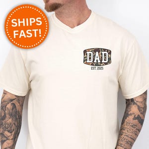 May include: Ivory-colored t-shirt with a V-neck. The shirt features a camouflage design with the word "DAD" and the text "EST. 2025". An orange circle with the words "SHIPS FAST!" is visible.