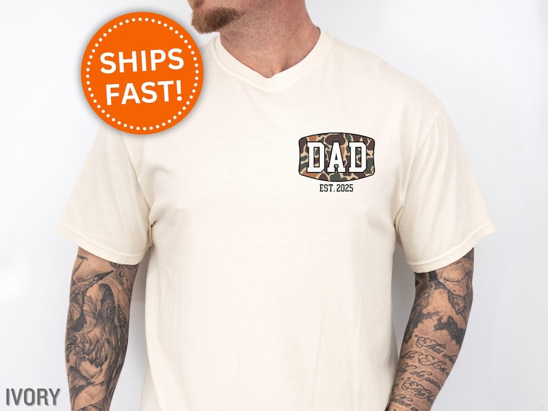 May include: Ivory-colored t-shirt with a V-neck. The shirt features a camouflage design with the word "DAD" and the text "EST. 2025". An orange circle with the words "SHIPS FAST!" is visible.