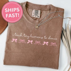 May include: Espresso-colored t-shirt with the phrase "i teach tiny humans to dance" in white script and pink bow graphics. A silver necklace with a pendant is on the shirt.