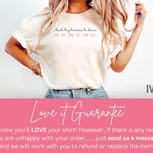 May include: Ivory-colored t-shirt with the text "i teach tiny humans to dance" in a script font. Below the text are pink bow graphics. The shirt is a casual style.