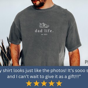 May include: A dark gray t-shirt with a white graphic of a sneaker and the text "dad life. est. 2025".