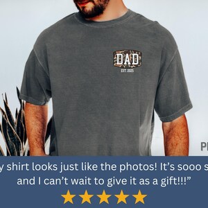May include: Dark gray t-shirt with a camouflage-style patch that reads "DAD EST. 2025". The shirt is a solid color and has short sleeves. The shirt is made of a soft material.