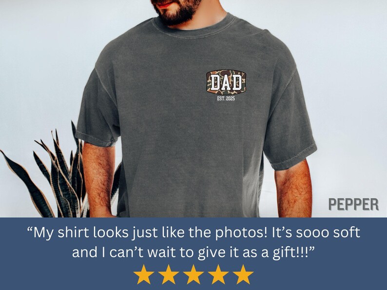 May include: Dark gray t-shirt with a camouflage-style patch that reads "DAD EST. 2025". The shirt is a solid color and has short sleeves. The shirt is made of a soft material.