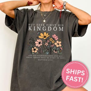 K&ouml;nnte beinhalten: Dunkelgraues T-Shirt mit dem Text "BUT SEEK FIRST HIS KINGDOM" &uuml;ber einem Blumendesign. Das Design umfasst rosa, gelbe und wei&szlig;e Blumen. Der Text "AND HIS RIGHTEOUSNESS AND ALL THESE THINGS WILL BE GIVET TO YOU. MATTHEW 6:33" befindet sich darunter.