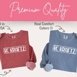 K&ouml;nnte beinhalten: Zwei Sweatshirts mit dem Aufdruck "KC KRAFTZ". Ein Sweatshirt ist staubig rosafarben und das andere hellblau.