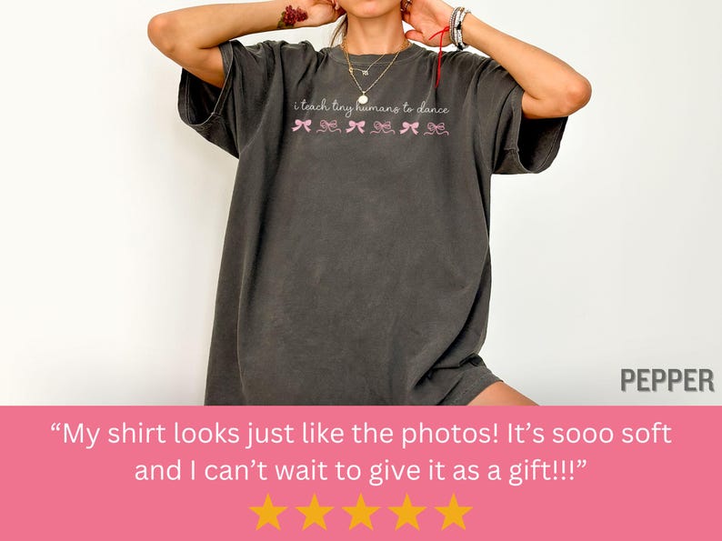 May include: Dark gray t-shirt with the text "i teach tiny humans to dance" in a script font, with pink bow graphics. The shirt is a relaxed fit, with short sleeves.