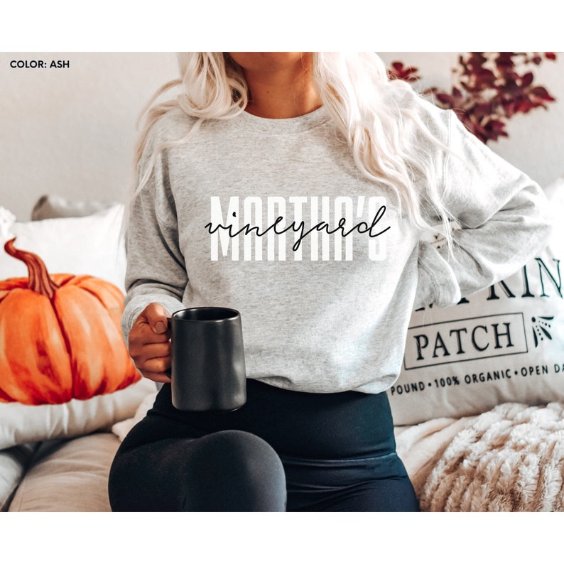 K&ouml;nnte beinhalten: Ein graues Sweatshirt mit dem Aufdruck "Martha's Vineyard" in Wei&szlig; auf der Vorderseite. Das Sweatshirt wird von einer Person getragen, die eine schwarze Tasse h&auml;lt.