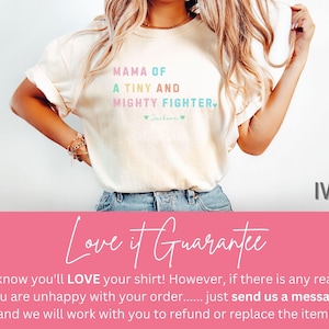 May include: Ivory-colored t-shirt with the text "MAMA OF A TINY AND MIGHTY FIGHTER" in pastel colors. The shirt has a crew neck and short sleeves. The word "Ivory" is printed on the right side of the shirt.