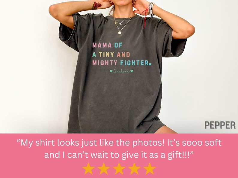 May include: Dark gray t-shirt with the text "MAMA OF A TINY AND MIGHTY FIGHTER" in colorful letters. The shirt also includes the name "Jackson" and two hearts. The shirt is a comfortable, oversized fit.