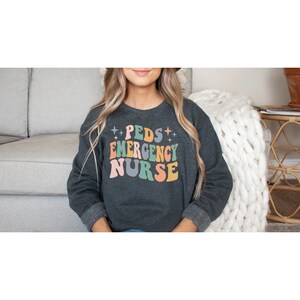 May include: A dark gray sweatshirt with the text "PEDS EMERGENCY NURSE" in colorful retro-style lettering. The text is surrounded by small white stars.